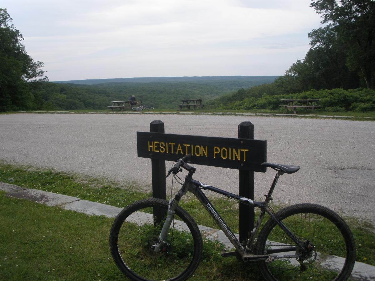 Gary Fisher Cobia: A mountain bike parked next to a sign that reads "Hesitation Point," with a scenic view of rolling hills and a few picnic tables in the background. A person is seated in the distance, enjoying the landscape under a cloudy sky.