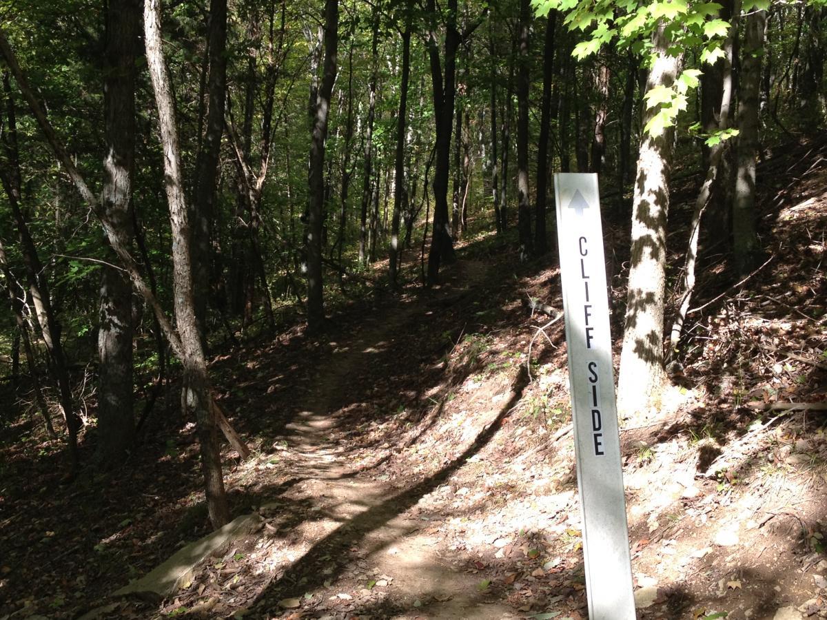 Pathway through a wooded area with a sign indicating "CLIFF SIDE," surrounded by trees and dappled sunlight filtering through the leaves. Versailles State Park mountain bike trail.