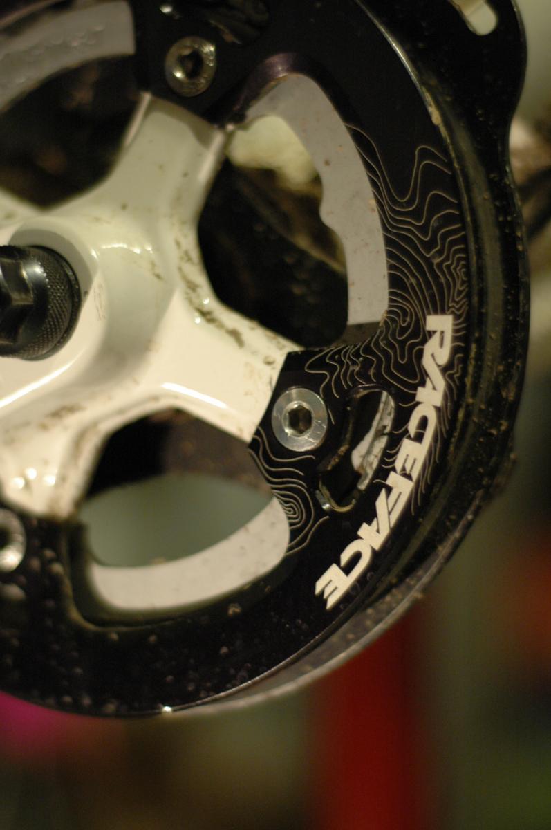 Race Face Lightweight Bash: Close-up of a bicycle crankset featuring a black chainring with a white contour design and the logo "RACEFACE." The chainring shows signs of dirt, indicating recent use. The background is blurred, with hints of a workshop environment.