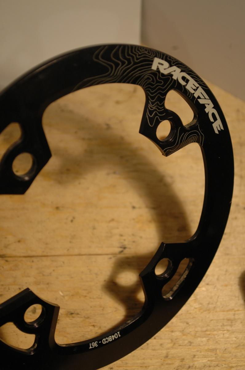 Race Face Lightweight Bash: Black bicycle chainring with the "RaceFace" logo and a design pattern, resting on a wooden surface. The chainring features multiple cutouts and is designed for enhanced performance in cycling.
