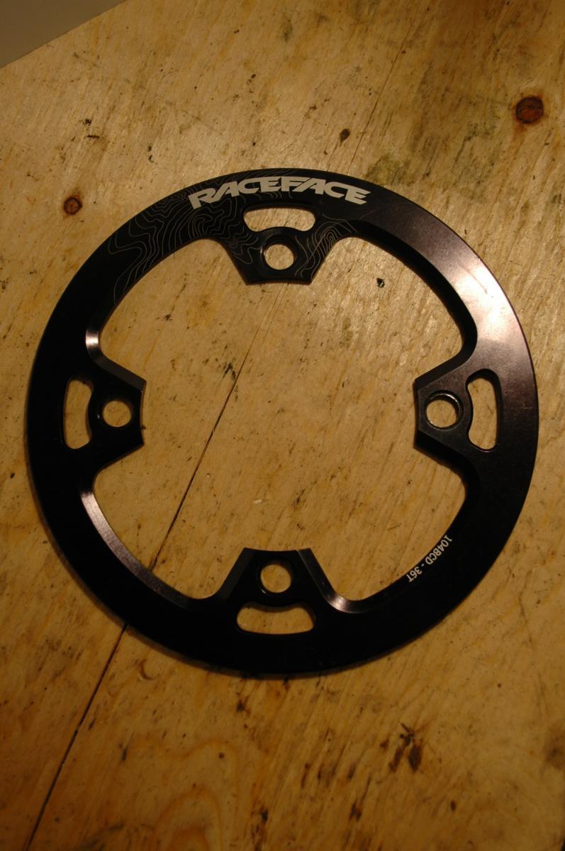 Race Face Lightweight Bash: Black bicycle chainring with a topographical design and multiple holes for mounting, resting on a wooden surface. The brand name "RACEFACE" is prominently displayed.