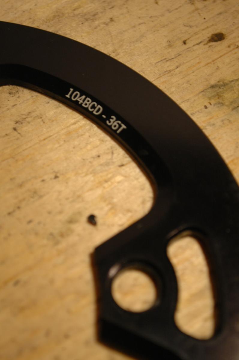 Race Face Lightweight Bash: Close-up image of a black bicycle chainring guard displaying the text "104BCD - 36T" on a textured wooden surface. The guard features a rounded shape with a cutout design for mounting.