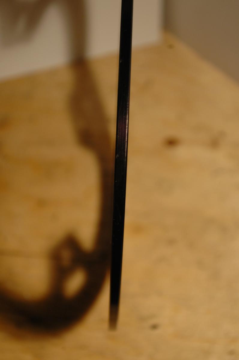 Race Face Lightweight Bash: A close-up image of a thin, vertical black object casting a shadow on a light wooden surface. The background is softly blurred, emphasizing the slim profile and clean edge of the object.