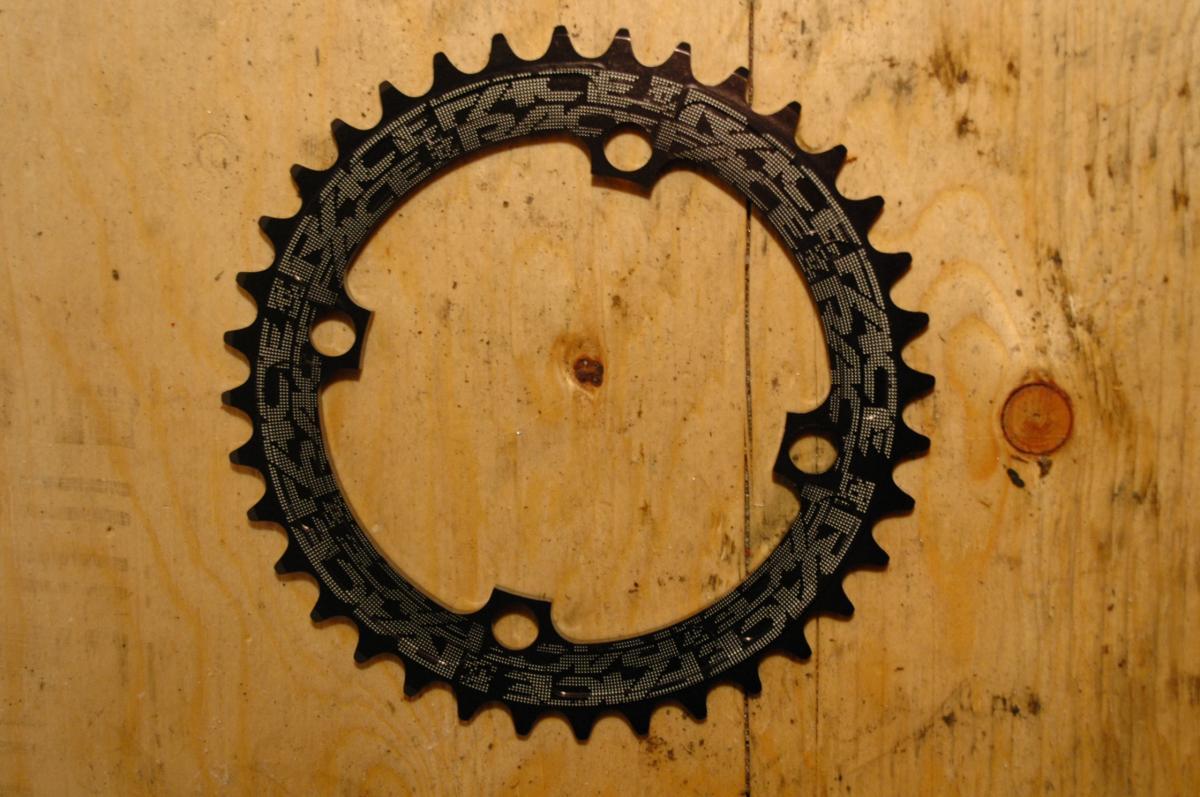 Race Face Single Chainring: Black bicycle chainring placed on a wooden surface, featuring a circular design with sharp teeth and multiple mounting holes, showing a textured pattern on its surface.