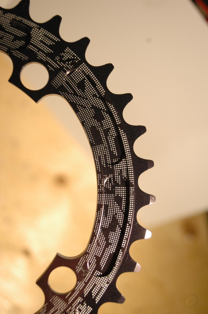 Race Face Single Chainring: A close-up image of a bicycle chainring with a black finish, featuring distinct teeth and detailed engravings on its surface. The background is softly blurred, highlighting the metallic sheen and intricate design of the chainring.