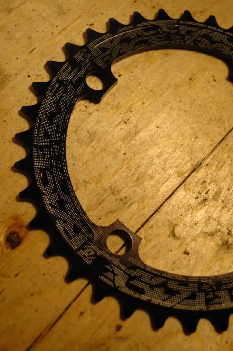 Race Face Single Chainring: A close-up image of a black bicycle chainring placed on a wooden surface, featuring a unique patterned design and several teeth along the edge. There are two holes for mounting, showcasing the intricate details of the component.