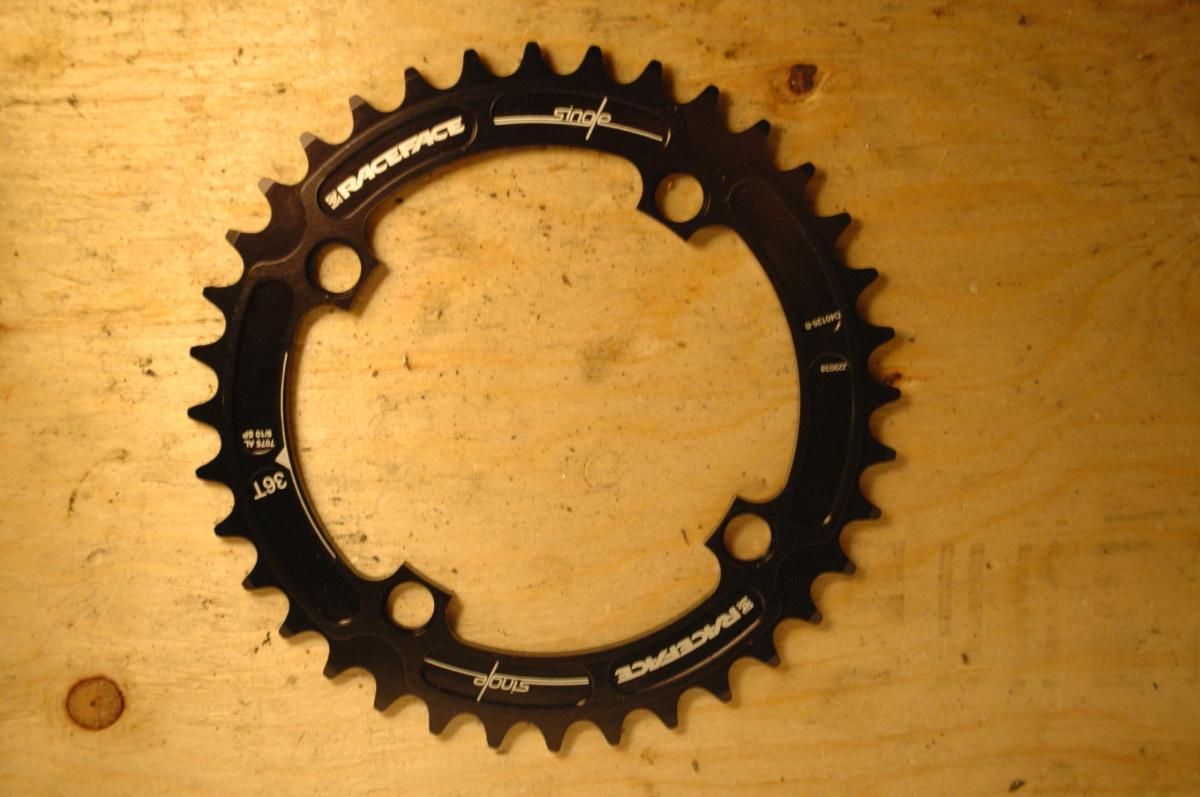 Race Face Single Chainring: Black bicycle chainring with teeth and mounting holes, placed on a wooden surface. The chainring features the brand name "Race Face" and is labeled with size markings.