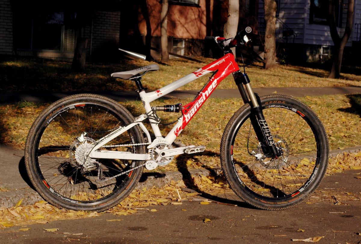Banshee Rampant: Mountain bike parked on a sidewalk surrounded by fallen leaves, featuring a white frame with red accents and high-performance tires. The bike has a prominent suspension fork and is showcased in a sunny autumn setting.