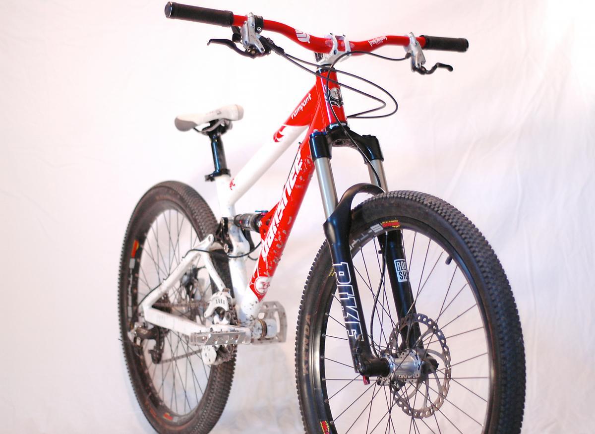 Banshee Rampant: A red and white mountain bike positioned at an angle, showcasing its front fork and handlebars. The bike features a robust frame, disc brakes, and knobby tires, set against a neutral background.