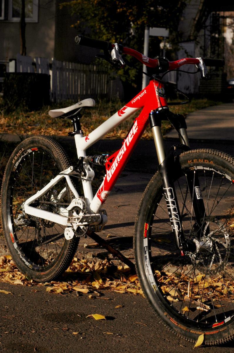 Banshee Rampant: A mountain bike with a white and red frame, featuring thick tires and a suspension system, parked on a leaf-strewn pavement. In the background, there are hints of a residential area with a white picket fence.