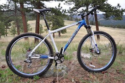 Redline D600: Mountain bike parked on rocky terrain surrounded by trees and a grassy field, with mountains in the background. The bike features a white and blue frame, thick tires, and a suspension fork.