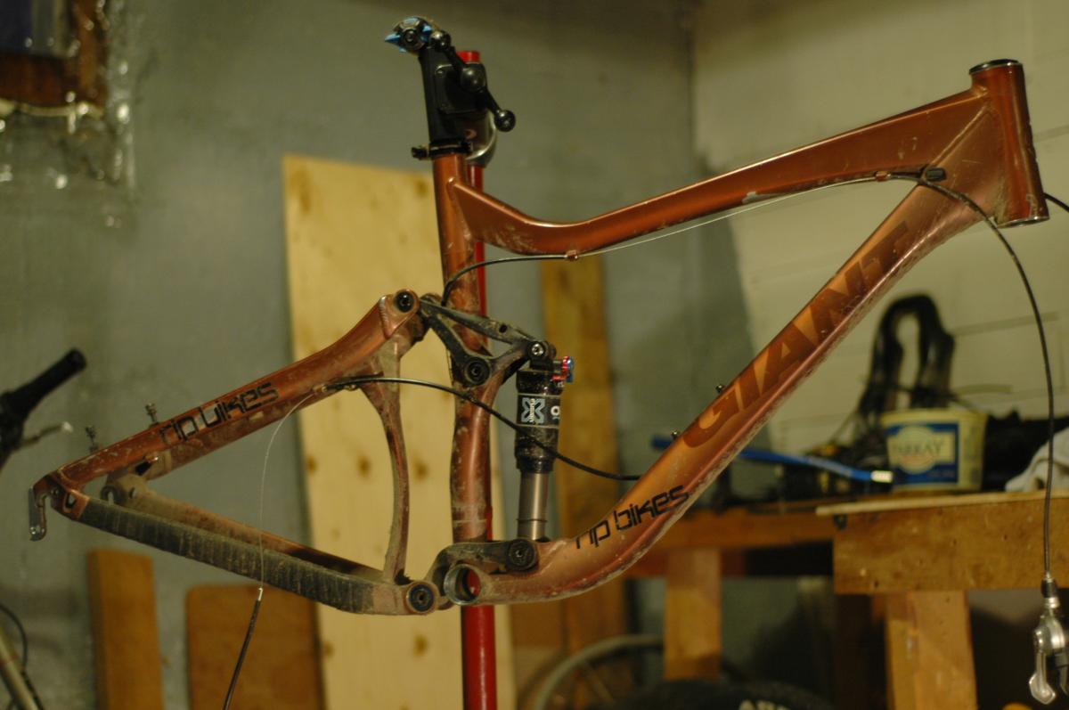 Giant Reign 1: A close-up image of a mountain bike frame, featuring a brown color with visible dirt, mounted on a repair stand. The frame has a suspension system and cables attached. In the background, there are wooden surfaces and bike tools, suggesting a workshop environment.