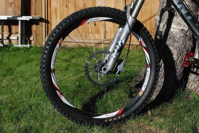 Giant Reign X1: A mountain bike wheel resting on grass, featuring a black tire with a patterned tread, a silver disc brake, and a lightweight aluminum fork. The background includes a wooden fence and a tree trunk, highlighting a bright, sunny day.