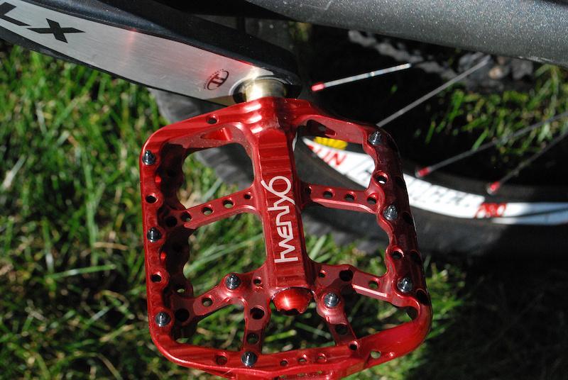 Giant Reign X1: Close-up image of a bright red bicycle pedal attached to a mountain bike, resting on green grass. The pedal features a unique design with multiple grip pins and the brand name "Twenty6" prominently displayed. A portion of the bike frame and tire are partially visible in the background.