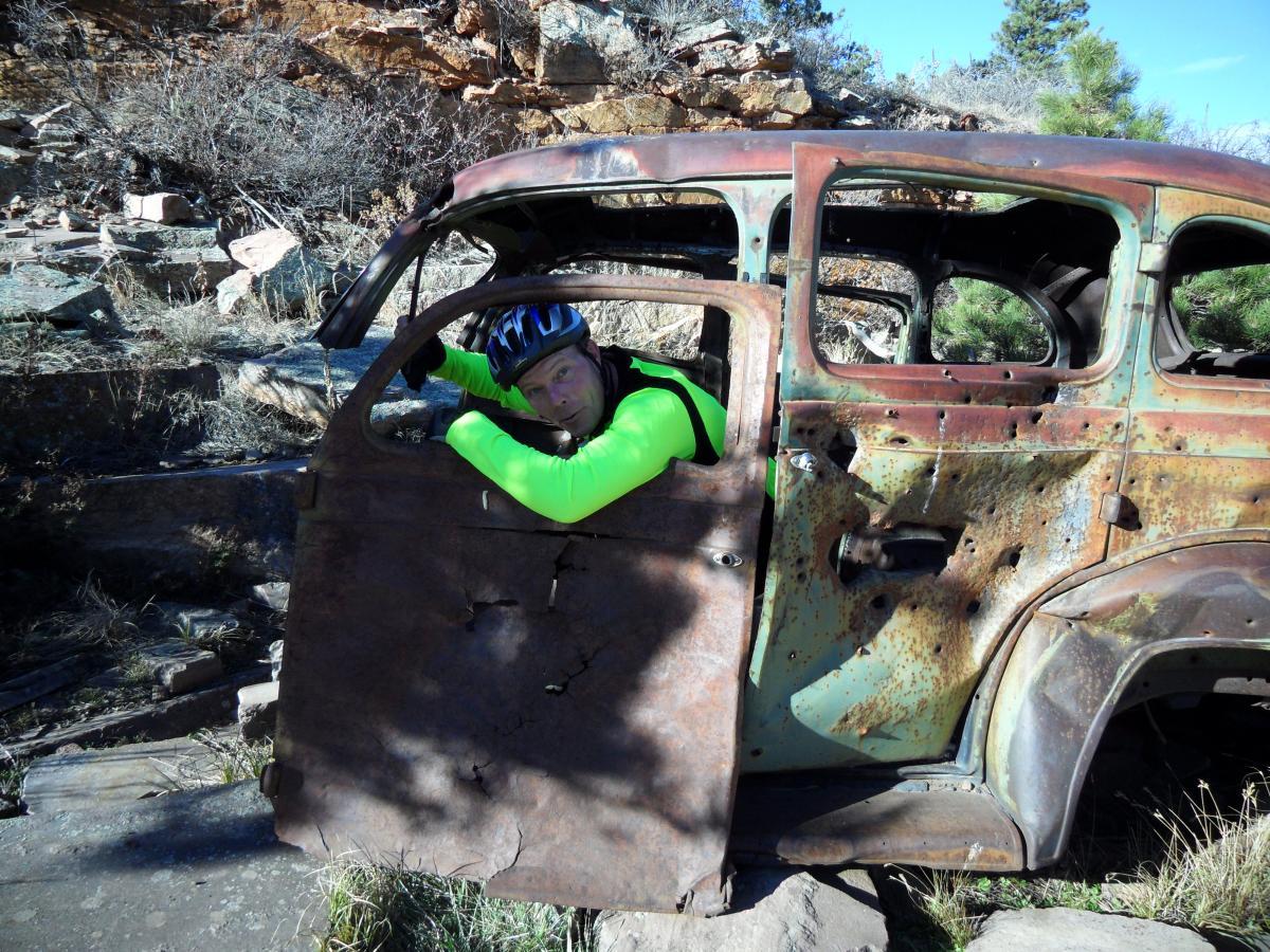 A person wearing a bright green long-sleeve shirt and a black helmet leans on the door of an old, rusted car in a rugged outdoor setting. The car, showing significant wear and decay, is surrounded by rocks and sparse vegetation under a clear blue sky. Picture Rock Trail mountain bike trail.
