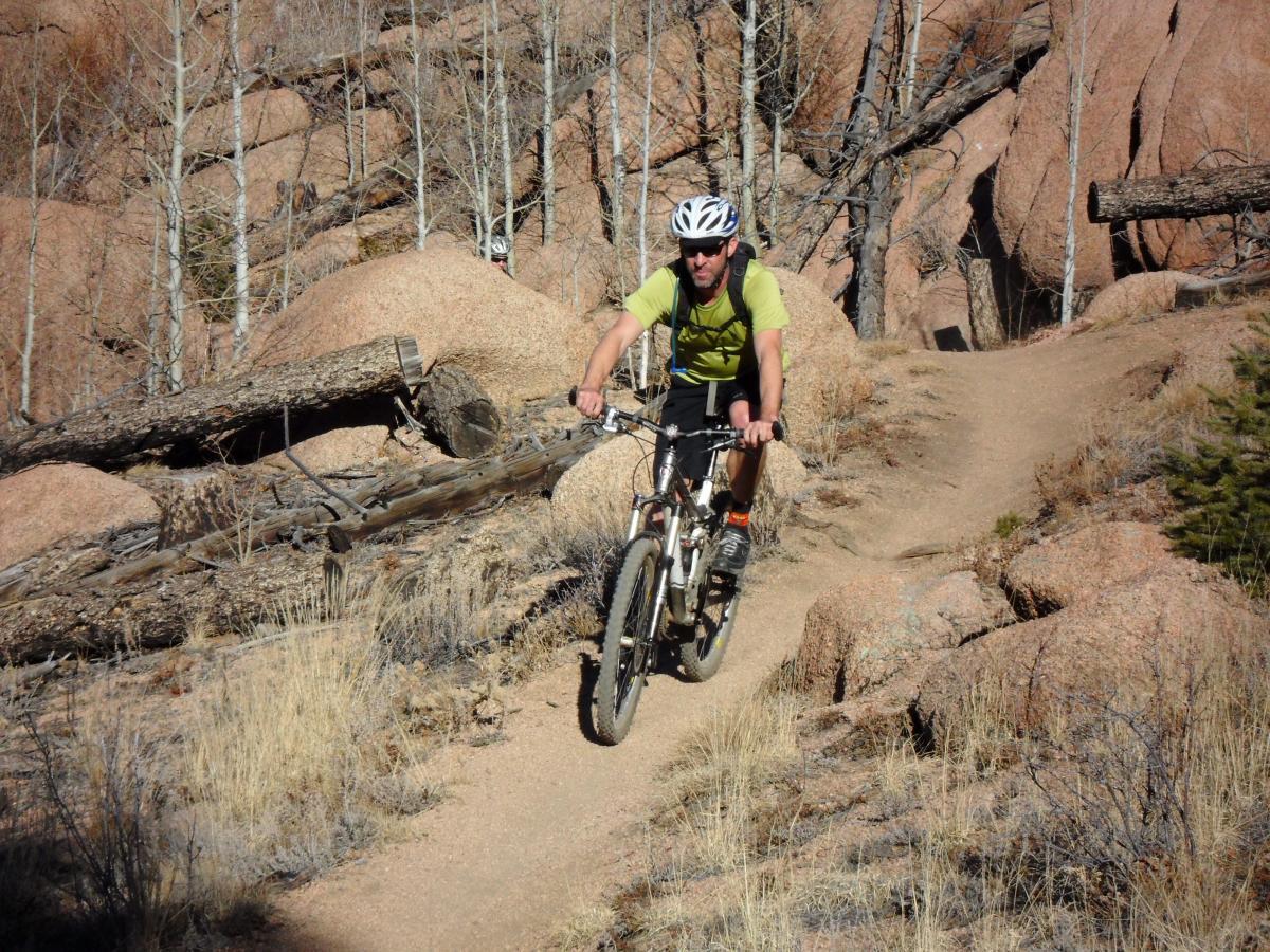 A cyclist navigating a rocky mountain bike trail surrounded by trees and boulders. The rider is wearing a helmet and a green shirt, showcasing dynamic movement as they maneuver along the path. Buffalo Creek mountain bike trail.