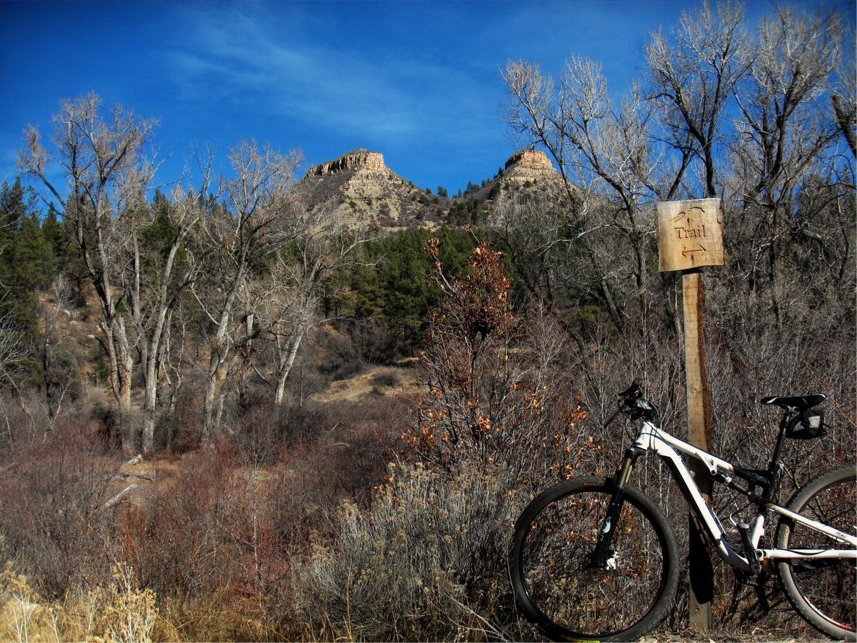 A mountain bike parked beside a wooden sign indicating a trail, with a backdrop of rocky formations and trees under a clear blue sky. Twin Buttes mountain bike trail.