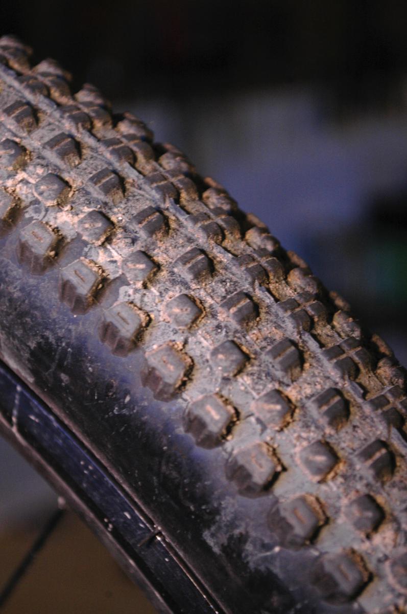 Kenda Small Block Eight: Close-up of a mountain bike tire showing rugged tread patterns and a layer of dirt, set against a blurred background.