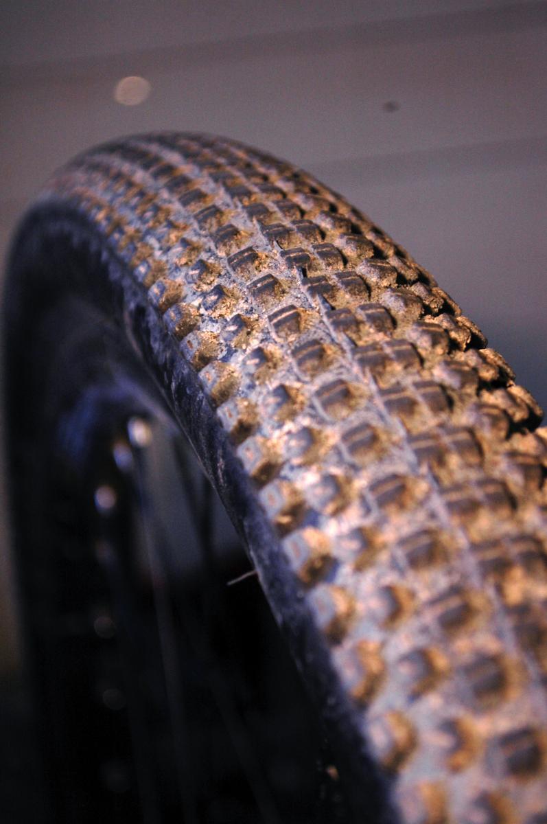 Kenda Small Block Eight: Close-up image of a worn bicycle tire with a textured tread pattern. The tire shows signs of dirt accumulation, indicating use on rough terrains. The background is softly blurred, focusing attention on the tire's detail.