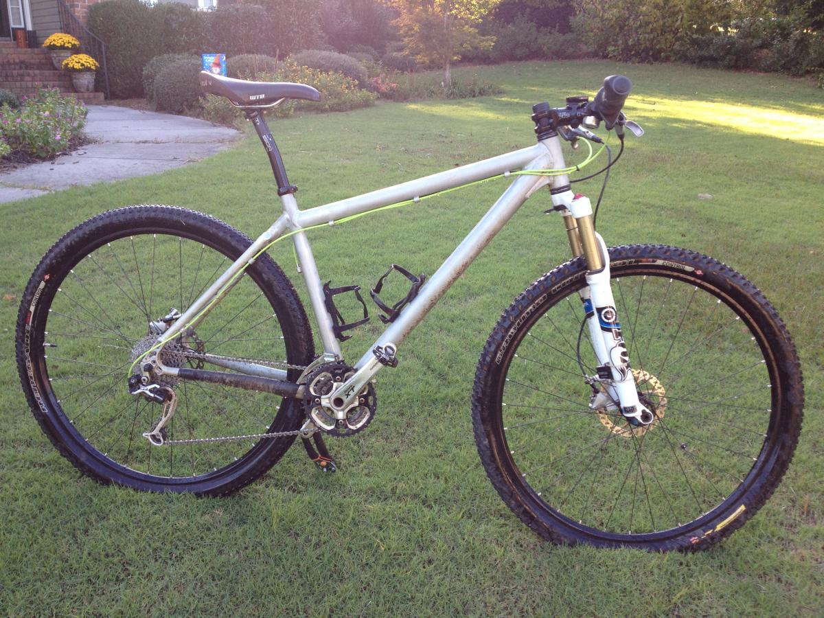 On-One Scandal 29er Vertical Dropouts: A silver mountain bike with knobby tires and a front suspension fork, parked on green grass. The bike features a single gear system and two water bottle holders, with a hint of dirt on the frame, indicating recent use. In the background, a landscaped area with shrubs and flowers is visible.