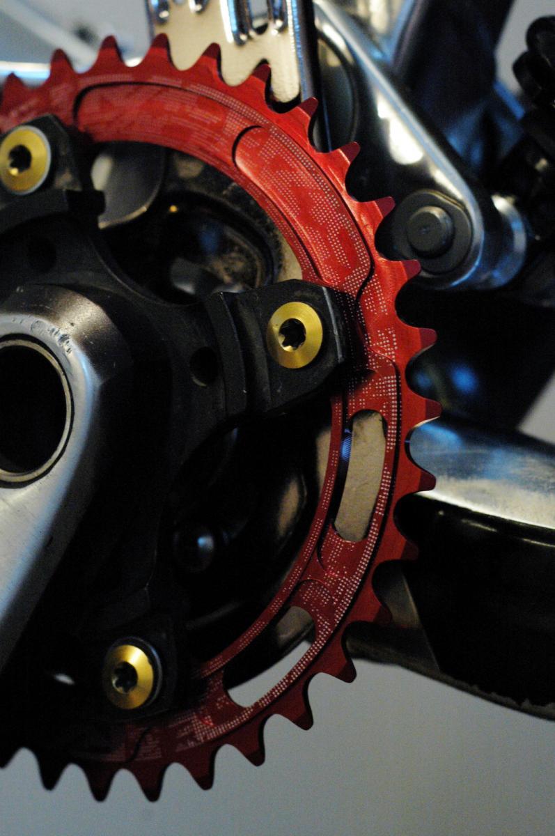 Race Face Single Chainring: Close-up image of a red bike chainring, showcasing detailed tooth design and installation on a black crankset. The background features metallic components, emphasizing the mechanical complexity of the bike.