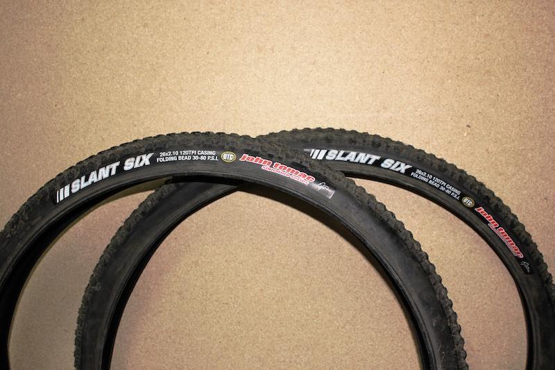 Kenda Slant Six: Two mountain bike tires labeled "Slant Six," resting on a textured surface. The tires have visible tread patterns and specifications, including the size 26 x 2.10 and a folding bead design.