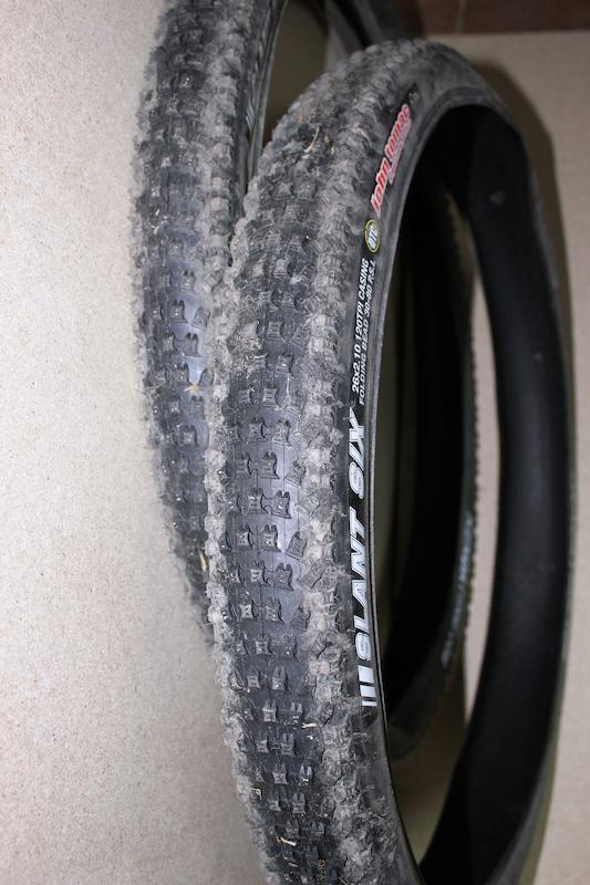 Kenda Slant Six: Two used mountain bike tires are leaning against a wall. The tires feature a knobby tread pattern for off-road traction and are partially covered in dirt, indicating previous use. The visible branding on the tire reads "Slant Six."