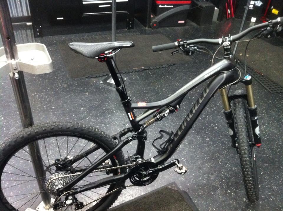 Specialized Stumpjumper FSR: A black mountain bike is positioned on a repair stand in a workshop. The bike features a suspension system, thick tires with knobby treads, and a sleek frame design. The background shows tools and equipment typically found in a bike maintenance area, with a dark flooring surface.