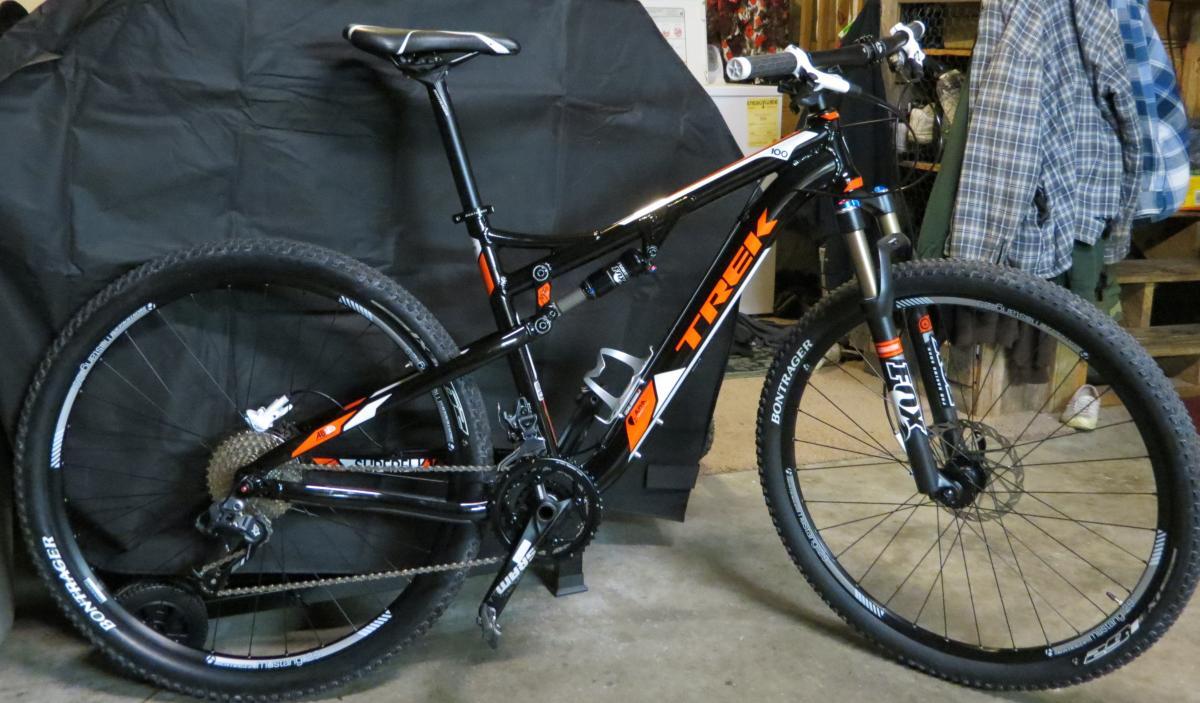 Trek Superfly: A black and orange Trek mountain bike is positioned against a dark background. The bike features thick tires, a dual-suspension frame, and several gear components, with a prominent "Trek" logo on the frame. In the background, various items are visible, including a plaid shirt hanging on a peg. The setting appears to be a garage or workshop.
