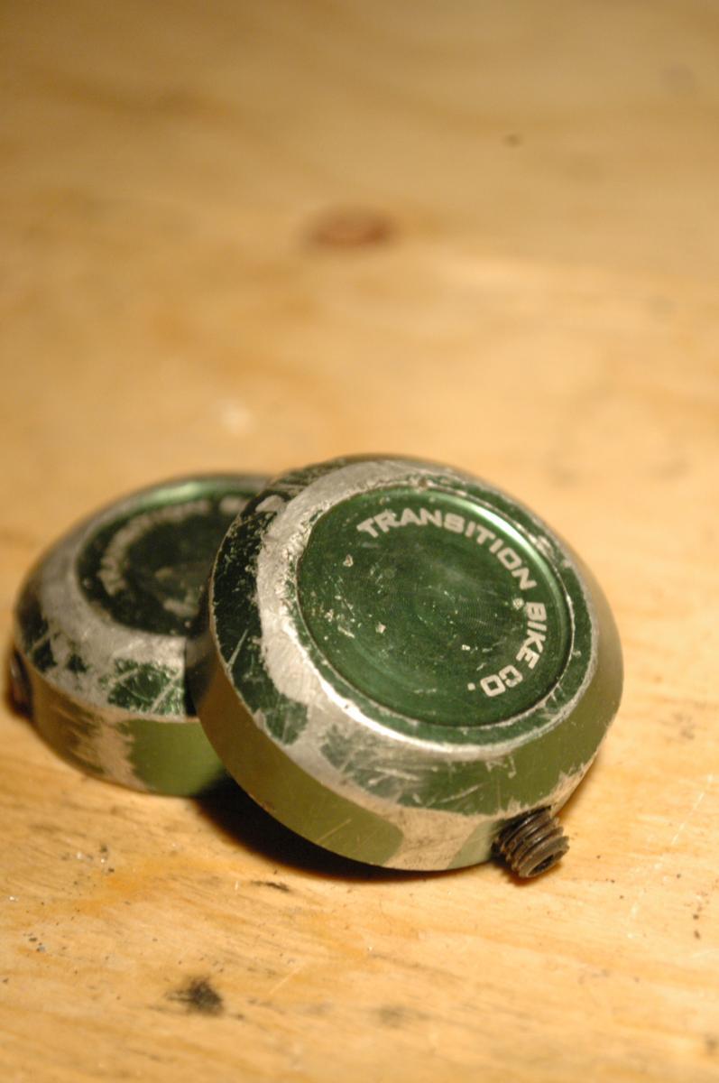 Transition Bar End Caps: Two green bike component caps are resting on a wooden surface. The caps show signs of wear with scratches and scuffs, and one features the text "Transition Bike Co." etched on it. The background is softly blurred, emphasizing the caps.