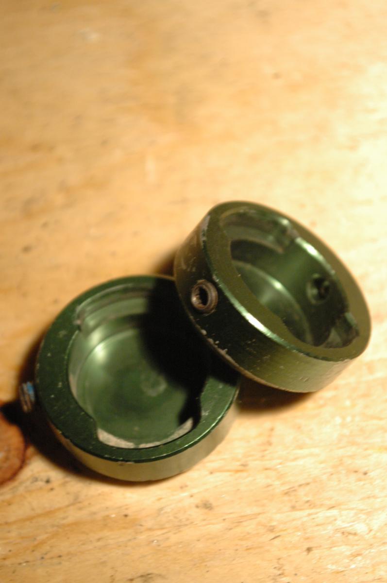 Transition Bar End Caps: Two green metal caps are shown, with one slightly atop the other, resting on a wooden surface. The caps are circular with a smooth finish and a small screw hole on one side. The lighting highlights their metallic texture and shine.