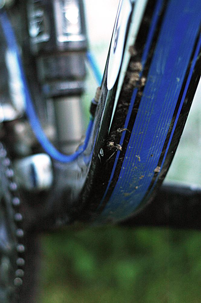 Giant Trance X2: Close-up view of a bicycle's frame and tire, highlighting a blue stripe along the tire and some dirt, suggesting recent outdoor use. The background is blurred, focusing on the details of the bike components.