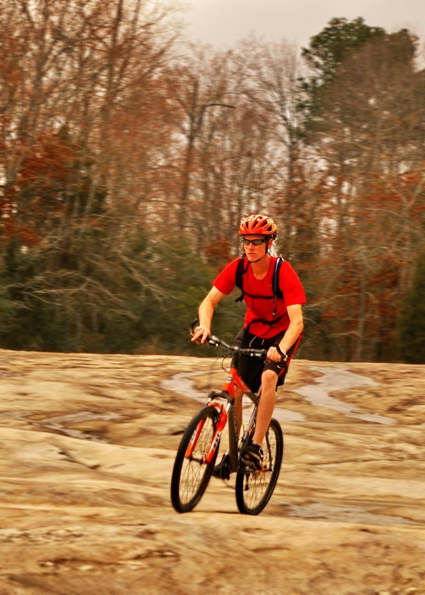 A cyclist in a red shirt and helmet rides a mountain bike over a rocky terrain. The background features a mix of bare and leafy trees, indicating a natural outdoor setting. Tribble Mill Park mountain bike trail.