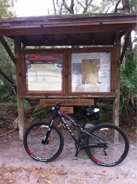 A mountain bike leaning against a wooden information kiosk in a natural outdoor setting. The kiosk features a map and informational signs about local trails and wildlife. Surrounding vegetation includes trees and shrubs, indicating a forested area. Spruce Creek Preserve mountain bike trail.