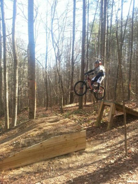 A mountain biker jumps off a wooden ramp in a forested area. The biker is mid-air, showcasing a dynamic leap, while trees surround the scene with sparse leaves. The ground is covered in brown foliage, indicating a natural trail setting. Freedom Park (aka:district Park) mountain bike trail.