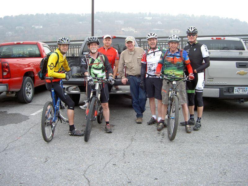A group of six mountain bikers poses for a photo near parked trucks. They are dressed in cycling gear and are holding their bikes, with a scenic background of hills in the distance. The mood is cheerful and the setting appears to be a parking lot, likely before a biking adventure. Burns Park mountain bike trail.