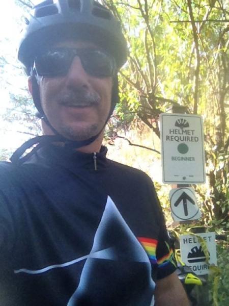 A person wearing a helmet and sunglasses smiles for the camera, dressed in a dark cycling shirt with a rainbow design. In the background, there are signs indicating that a helmet is required and that the trail is suitable for beginners. The surrounding area is lush with greenery. Amelia Earhart Park mountain bike trail.