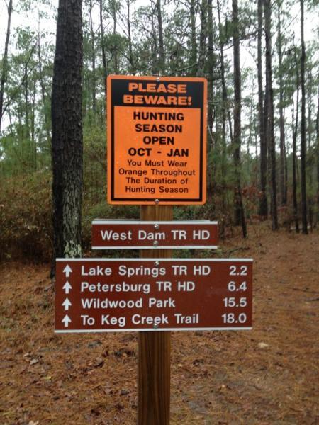 A sign in a forest area indicating that hunting season is open from October to January. It warns visitors to wear orange for safety. Below, there’s a directional sign for trails, including distances to Lake Springs, Petersburg, Wildwood Park, and Keg Creek Trail. The area is surrounded by tall trees, suggesting a wooded environment. Bartram Trail / West Dam / Wildwood Park mountain bike trail.
