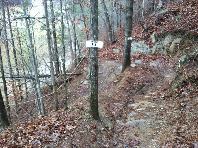 A narrow, winding dirt path surrounded by trees, with signs reading "SLOW!" on two posts. The ground is covered with dried leaves and there are visible rocks along the trail. A slight incline is present as the path leads through a wooded area. Van Michael Trail mountain bike trail.