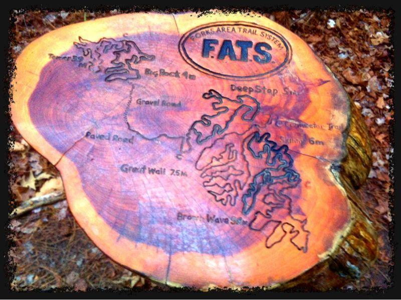 A carved wooden sign depicting a map of the Forks Area Trail System (FATS). The sign features various trail names and distances, outlined in dark engraving on the wood. The top of the sign includes the FATS logo, and the surrounding area is covered in fallen leaves, suggesting a natural outdoor setting. Forks Area Trail System (FATS) mountain bike trail.