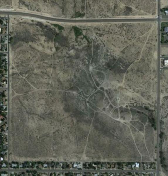 Aerial view of a sparsely vegetated landscape featuring dirt paths winding through barren terrain, with residential neighborhoods visible along the bottom edge of the image. The Hills mountain bike trail.