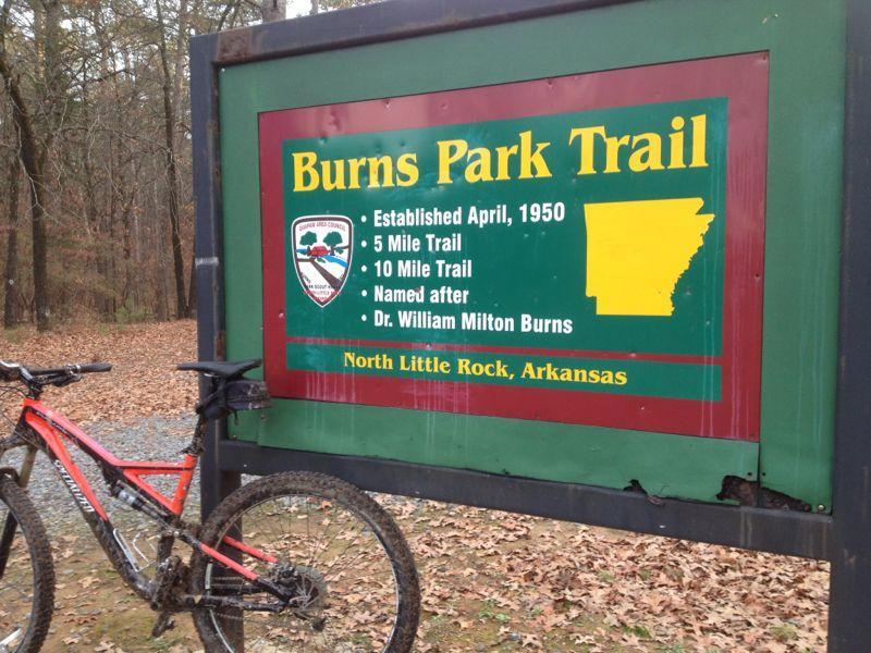 Sign for Burns Park Trail in North Little Rock, Arkansas, detailing trail information including established date, trail lengths, and the person it is named after, alongside a parked mountain bike. Burns Park mountain bike trail.