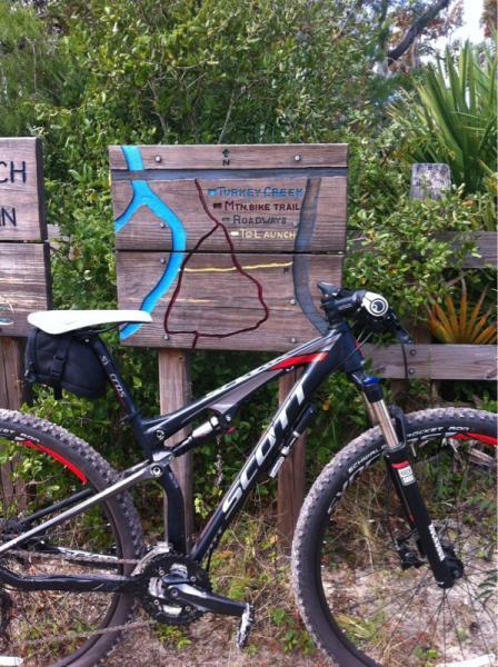 A mountain bike leaning against a wooden sign displaying a map of the Tamiami Creek Mountain Bike Trail, showing routes and directions. Surrounding vegetation is visible, adding to the outdoor setting. Turkey Creek mountain bike trail.