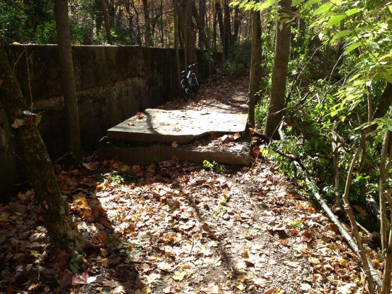 A narrow wooded path covered with autumn leaves, flanked by trees. A concrete slab lies along the path, partially eroded, with a bicycle parked nearby against a low stone wall. Sunlight filters through the tree canopy, creating patches of light and shadow on the ground. Fort Harrison State Park mountain bike trail.