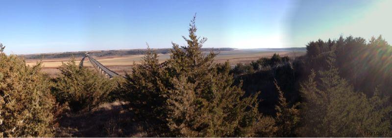 A panoramic view of a landscape featuring rolling hills and a river below, framed by lush green trees in the foreground. The sky is clear with a hint of sunlight illuminating the scene, creating a serene and tranquil atmosphere. Fancy Creek State Park mountain bike trail.