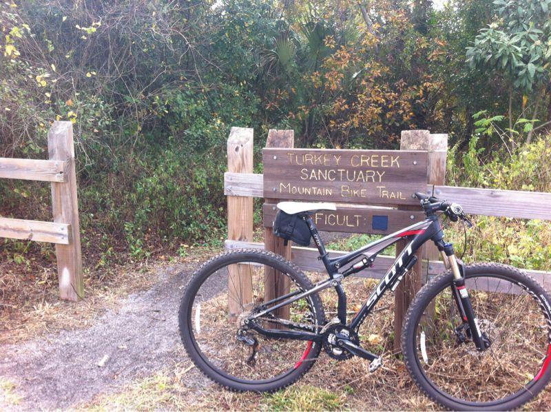 A mountain bike leaning against a wooden sign that marks the entrance to the Turkey Creek Sanctuary Mountain Bike Trail, labeled as "Difficult". The surrounding area features greenery and natural foliage. Turkey Creek mountain bike trail.