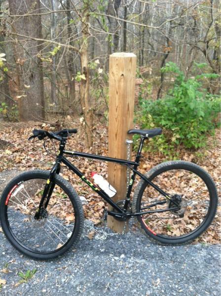 A black mountain bike parked next to a wooden post in a wooded area. The ground is covered with fallen leaves, and there are sparse trees in the background, indicating a natural trail. The bike features a water bottle mounted on the frame and has a rugged design suitable for outdoor cycling. Cabin John Trail mountain bike trail.