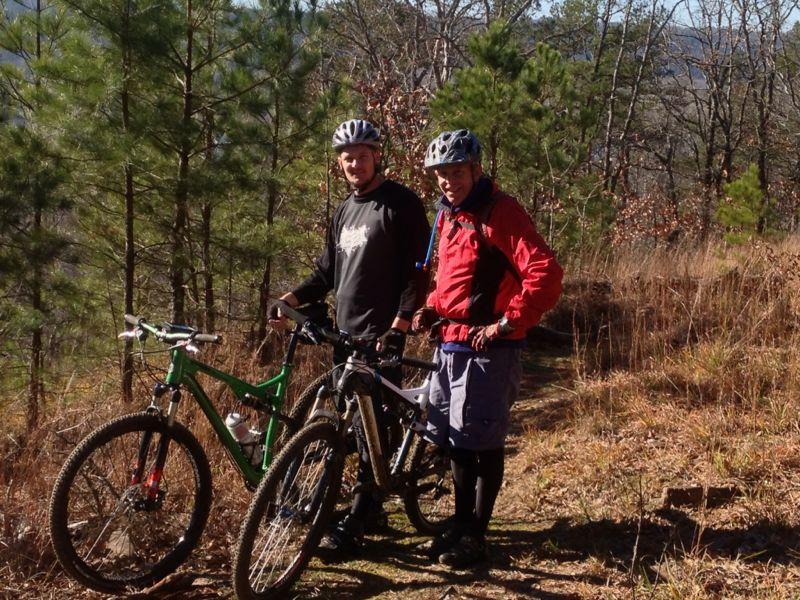 Two mountain bikers posing with their bicycles on a trail surrounded by trees. One rider is wearing a black shirt and a helmet, while the other is dressed in a red jacket with a hydration pack. Sunlight filters through the trees, creating a bright and inviting outdoor scene. Burns Park mountain bike trail.