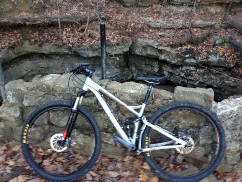 A mountain bike parked on a rocky ledge near a deep ravine, surrounded by fallen leaves and autumn vegetation. A wooden fence is visible in the background. Monte Sano State Park &amp; Land Trust mountain bike trail.
