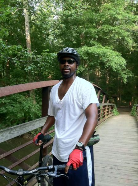A person wearing a black helmet and sunglasses stands beside a bicycle on a wooden bridge surrounded by lush green trees. The individual is dressed in a white t-shirt and dark shorts, appearing to have been actively cycling. Wakefield mountain bike trail.